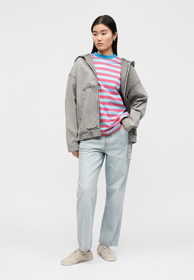 Νεαρή γυναίκα που φορά γκρι oversized μπουφάν, ροζ και μπλε ριγέ πουκάμισο, ανοιχτό μπλε τζιν και μπεζ αθλητικά παπούτσια, στέκεται με το ένα χέρι στην τσέπη.