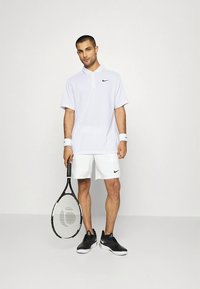 Wit poloshirt met een zwart Nike-logo, gecombineerd met witte short. Het model houdt een tennisracket vast en draagt zwarte sportschoenen.