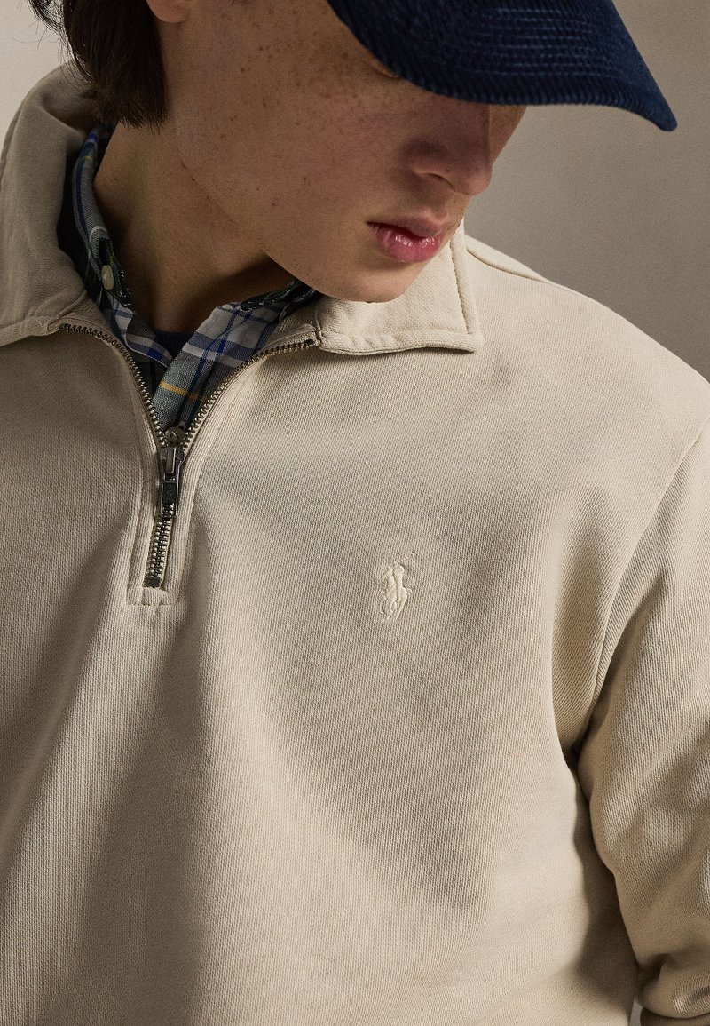 Young person wearing beige quarter-zip sweater over blue plaid shirt and dark blue cap, looking downward, neutral background.