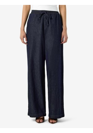 Femme portant un pantalon large bleu foncé avec taille élastique et cordon, associé à des sandales marron à bout ouvert et un haut blanc.