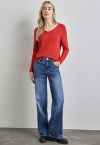 Rode v-hals trui met lange mouwen, gecombineerd met blauwe wijde jeans. De jeans hebben een knoopsluiting en voorkruiden.