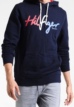 Person trägt einen dunkelblauen Kapuzenpullover mit rotem und hellblauem "Hilfiger Denim" Text und hellgrauen Jeans, gezeigt vom Hals bis zur Hüfte.