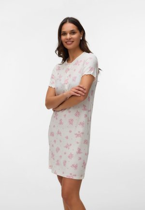Donna sorridente con le braccia incrociate, indossa un vestito bianco con stampa floreale rosa e maniche corte arricciate, su sfondo bianco uniforme.