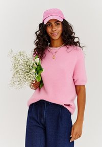 Krátky ružový načechraný sveter s krátkymi rukávmi nosený cez tmavomodré rifle s vysokým pásom. Drží kyticu bielych kvetov. Na hlave má ružovú čiapku.