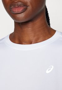 Biała sportowa koszulka z okrągłym dekoltem i krótkimi rękawami, z małym odblaskowym logo na piersi. Gładki, elastyczny materiał.