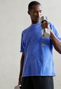 Man in blue shirt lifting a silver dumbbell with one arm, looking at the weight, against a plain light background.