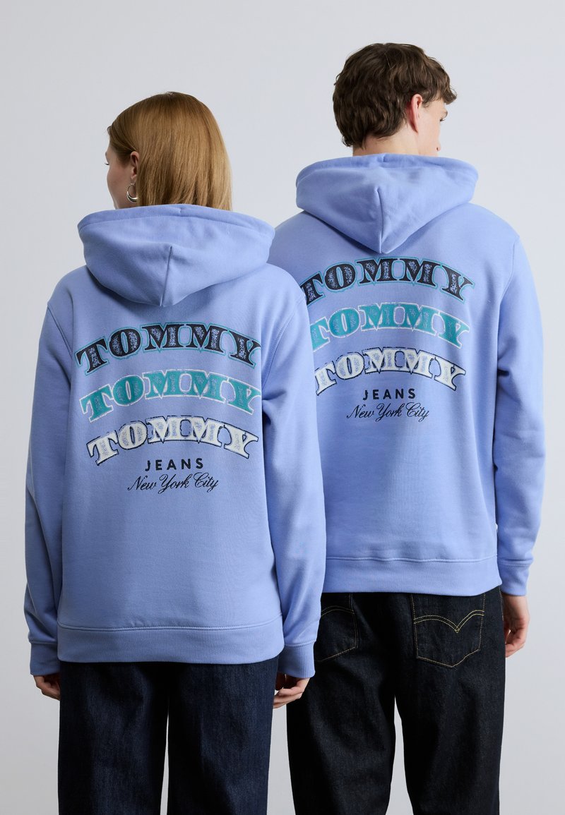 Hellblaue Kapuzenpullover mit dem Wort "TOMMY" in verschiedenen Schriftarten und Farben auf dem Rücken. Gerippte Bündchen und Saum, getragen mit dunkler Hose.