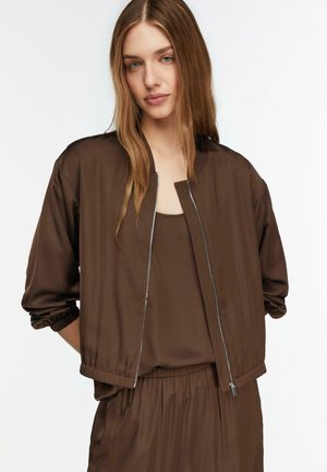 Młoda kobieta z długimi jasnobrązowymi włosami ma na sobie brązową satynową kurtkę bomber, top i pasujące spodnie z elastycznym stanem, pozując na białym tle.