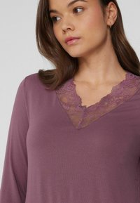 Kobieta w mauve'owej bluzce z długim rękawem i dekoltem w serek wykończonym koronką, patrzy w bok na jednolity jasny tle.