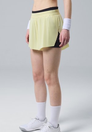 Personne portant une jupe de sport plissée jaune pâle avec des shorts noirs en dessous, des bracelets blancs aux poignets, des chaussettes blanches et des chaussures de sport blanches.