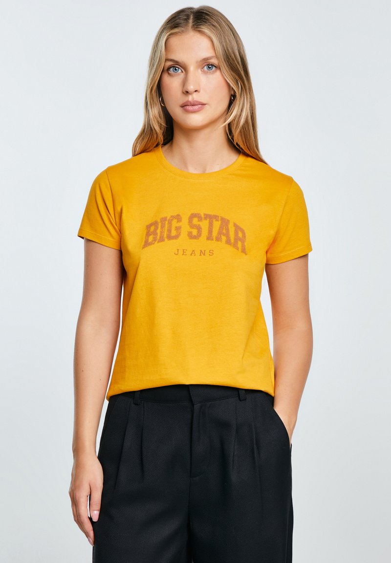 Geltona medvilninė marškinė su trumpomis rankovėmis ir apvaliu iškirpimu, su tamsiai raudonai parašytu "BIG STAR JEANS" tekstu krūtinės srityje.