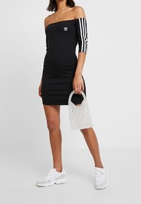 Svart off-the-shoulder klänning med trekvarts randiga ärmar, Adidas-logga och en vit vävd handväska, i kombination med vita sneakers.