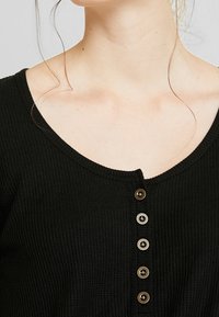 Schwarzes, geripptes Shirt mit weiter Ausschnittöffnung, versehen mit fünf runden Knöpfen in einer Reihe, und feiner Textur auf dem Stoff.