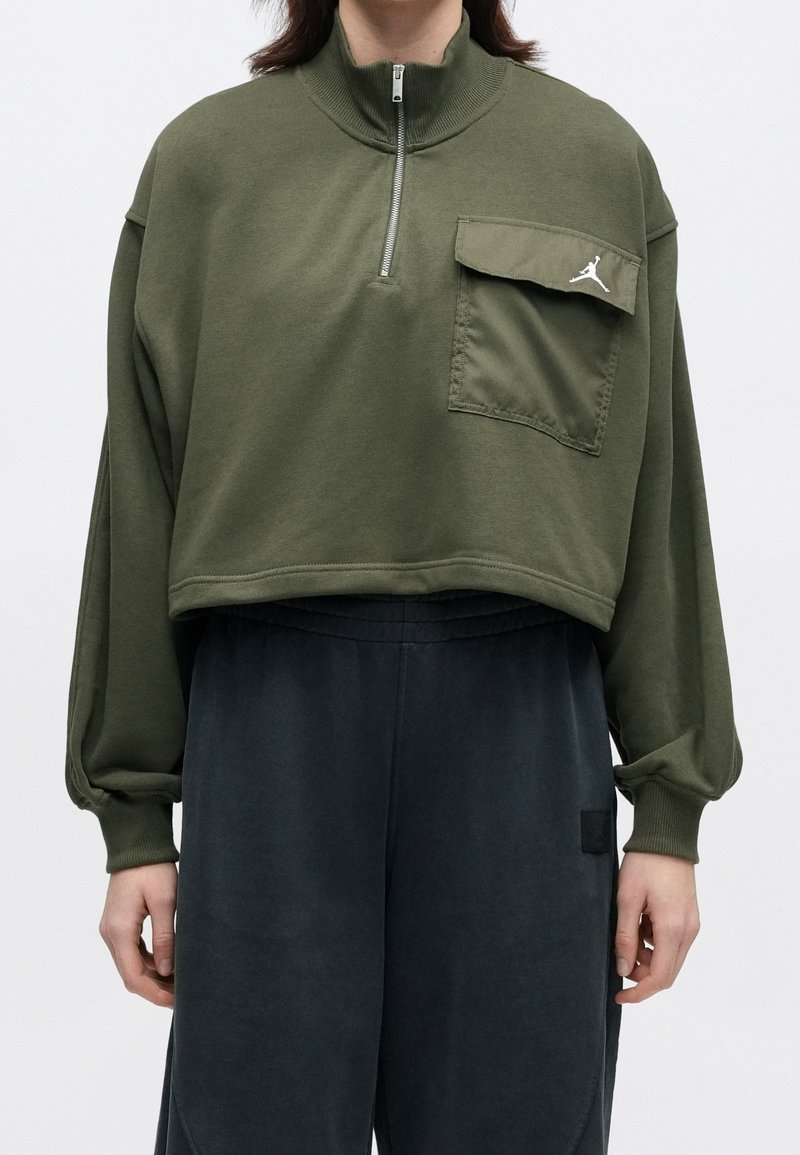 Personne portant un pull court vert olive avec une poche poitrine et un demi-zip, assorti à un pantalon sombre et large, debout devant un fond uni.