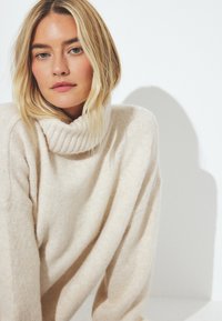 Hell-beige Rollkragenpullover aus weichem, strukturiertem Stoff. Mit weiten Ärmeln und einem lockeren Sitz. Einfaches und minimalistisches Design.