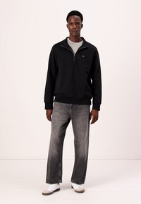 Felpa nera con zip e orlo a costine, abbinata a jeans larghi grigi e sneakers bianche. Design casual con un logo discreto sul petto.