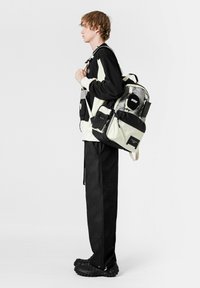 La mochila presenta una combinación de colores negros y claros, con un tejido texturizado, múltiples bolsillos y correas ajustables para opciones de transporte versátiles.