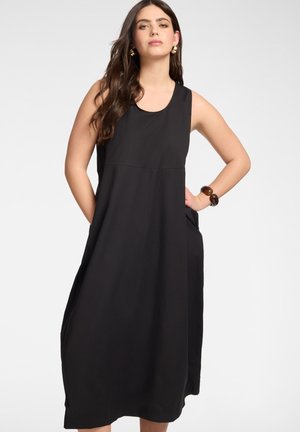 Frau trägt ein ärmelloses lockeres schwarzes Midikleid mit Taschen, goldene Creolen und ein grobes braunes Armband, steht mit den Händen in den Taschen.