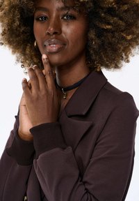 Bruine blazer met een gestructureerde kraag en geribbelde manchetten. De model heeft krullend haar en houdt haar handen bij haar gezicht, terwijl ze een zwarte choker draagt.