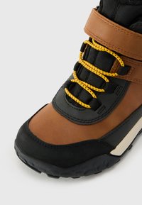 Botas de senderismo marrones y negras con una suela de goma texturizada, cordones amarillos y un cuello acolchado, que cuentan con una correa de seguridad en el tobillo.