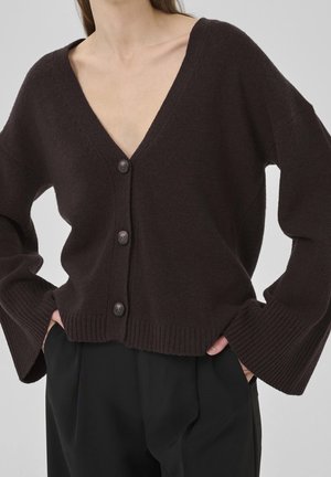 Cardigan marron foncé boutonné avec de larges manches porté sur un pantalon noir, montré du torse jusqu’aux hanches.