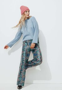 Un pull en maille bleu clair associé à un pantalon paillettes à jambes larges orné de motifs floraux verts et roses. Le mannequin porte un bonnet rose.