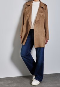 Persona vestida con un abrigo de ante color beige sobre una blusa blanca, jeans de pierna ancha azul oscuro y zapatillas blancas, de pie contra una pared gris clara.