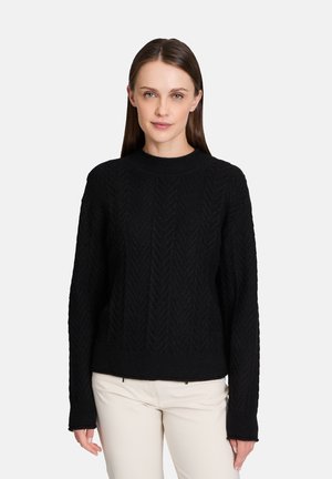 Zwarte gebreide trui met een textuur herringbone patroon, ronde halslijn en lange mouwen, gecombineerd met lichtgekleurde broek.