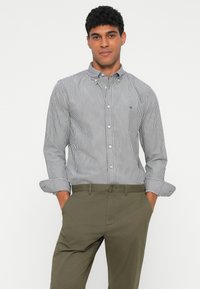Striped button-up shirt in navy and white, featuring a collar and rolled cuffs, paired with olive green trousers, with a small logo on the chest.