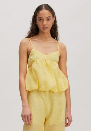 Femme aux cheveux ondulés portant des boucles d'oreilles spirales dorées, un top sans manches jaune texturé et un pantalon taille haute assorti, debout sur un fond neutre.