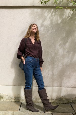 Femme souriante appuyée contre un mur clair texturé, portant un chemisier foncé, un jean bleu, des bottes marron jusqu'aux genoux et tenant un petit sac noir.