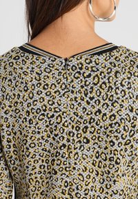 Haut à motifs noir, jaune et gris avec une fermeture éclair dans le dos et un col rayé. Présente un tissu texturé avec un motif léopard.