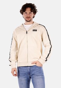 Felpa beige con zip frontale, polsini a coste e tasche laterali. Logo nero sul petto e bande laterali nere a contrasto. Texture morbida.