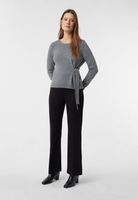 Frau steht mit den Händen hinter dem Rücken, trägt einen grauen Pullover mit langen Ärmeln und Seitenschleife, schwarze Hose und schwarze Stiefeletten mit Absatz.