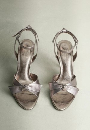 Sandali con tacco alto in argento metallico, dotati di una fascia anteriore annodata e di una fascia alla caviglia, che mostrano una texture liscia e lucida e un design con punta aperta.