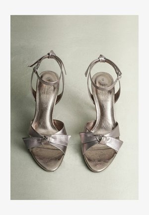Metāliskās sudraba augstpapēžu sandales ar mezglotu priekšējo siksniņu un potītes siksniņu, demonstrējot gludu, spīdīgu tekstūru un atvērtu purngalu.