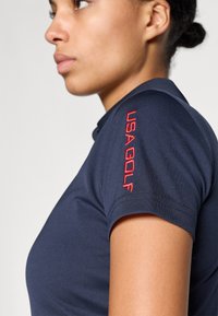 Tumši zils polo krekls ar pieguļošu dizainu, īsām piedurknēm un sarkanu "USA GOLF" izšuvumu uz kreisās piedurknes. Gluda auduma tekstūra.