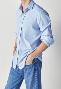 Vaaleansininen napillinen paita, jossa on pehmeä rakenne, pitkät hihat käännettynä cuffeihin, ja klassinen kaulus, yhdistettynä sinisiin farkkuihin.