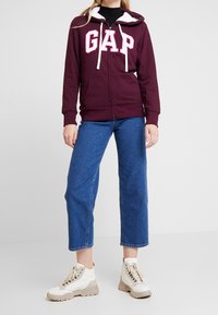 Personne portant un sweat à capuche zippé GAP bordeaux avec doublure en polaire blanche, jean bleu raccourci et baskets blanches épaisses, debout sur un fond blanc.