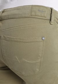 Jeans de algodón beige con un bolsillo trasero que presenta una costura curvada y una pequeña placa de logo. La tela tiene una textura suave y mate.
