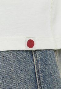 White t-shirt hem with a textured tag featuring a red dot, above blue denim fabric, highlighting a casual, layered look.