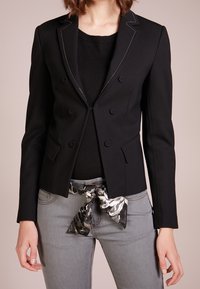 Černé dvouřadové sako s bílým prošíváním, s klopami a dvěma kapsami; nošeno přes černý top a šedé džíny s vzorovaným opaskem.