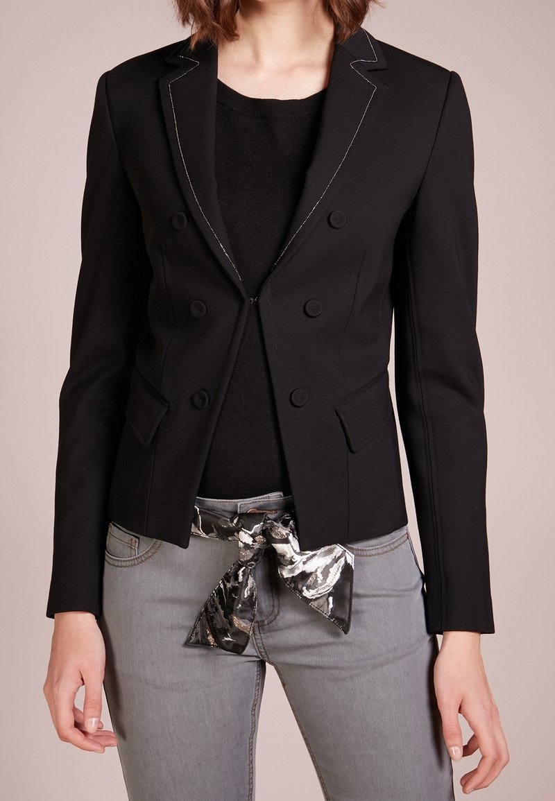 Černé dvouřadové sako s bílým prošíváním, s klopami a dvěma kapsami; nošeno přes černý top a šedé džíny s vzorovaným opaskem.