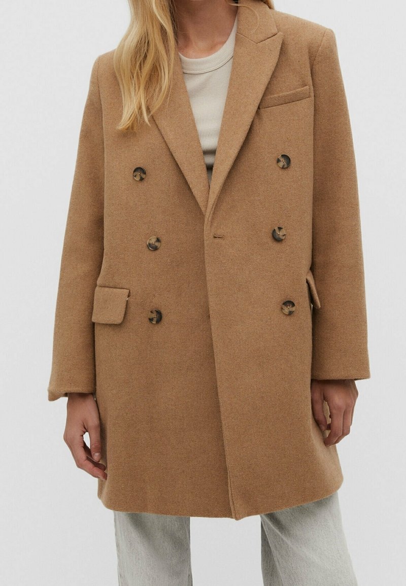Femme portant un manteau camel en laine à double boutonnage avec six boutons, deux poches latérales et une poche poitrine, assorti à un pantalon gris clair.