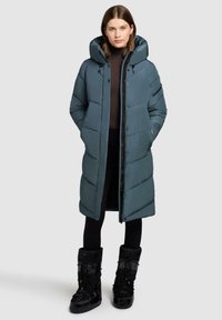 Kvinde iført en lang, teal-farvet, polstret vinterjakke med hætte, sorte leggings og sorte vinterstøvler, stående mod en ensfarvet baggrund.