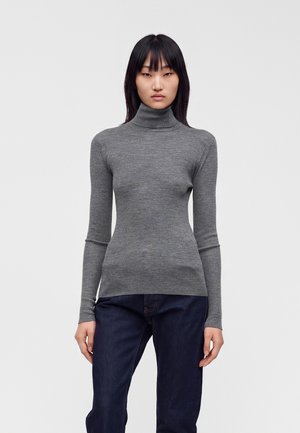Vrouw met steil zwart haar, gekleed in een nauwsluitende grijze geribbelde coltrui en donkerblauwe spijkerbroek, staand tegen een effen lichte achtergrond.