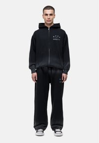 Zwarte zip-up hoodie met een voorzak en graphic tekst, gecombineerd met bijpassende zwart losvallende sweatpants en sneakers.