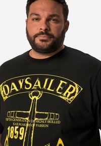 Černé tričko s žlutým grafickým potiskem kotev a slova "DAYSAILER." Materiál vypadá měkký, s kulatým výstřihem a krátkými rukávy.