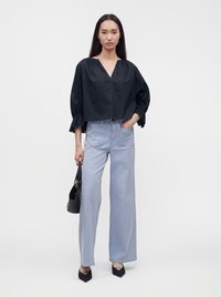 Femme aux longs cheveux foncés portant un chemisier noir ample et un jean large bleu clair, tenant un sac à main noir dans une main.