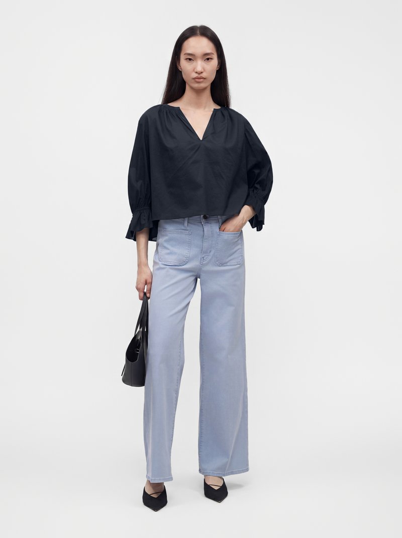 Femme aux longs cheveux foncés portant un chemisier noir ample et un jean large bleu clair, tenant un sac à main noir dans une main.